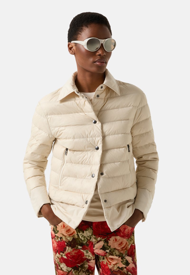 Světle béžová prošívaná bunda s límcem, vodorovným prošitím, dvěma kapsami na zip, doplněná kalhotami s květinovým vzorem a slunečními brýlemi.
