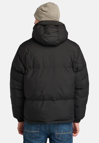 Sort puffet jakke med hætte, udstyret med et quiltet design og elastiske manchetter, bæres over blå jeans og ledsages af en lys grå hue.