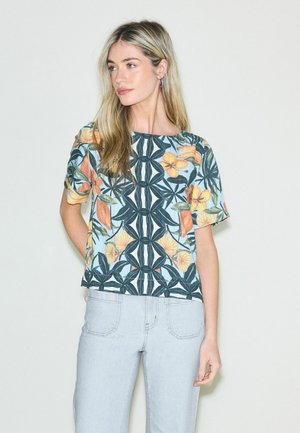 Vrouw met lang blond haar, gekleed in een korte mouwen top met bloemenpatroon en lichtblauwe spijkerbroek, staande tegen een effen lichte achtergrond.