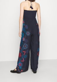 Marinblå jumpsuit med en figursydd smockad bodice och vida byxor. Har färgglada blommönster i röd och blå. Flätade band med tofsar.