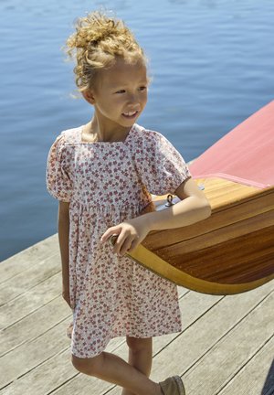 Light pink floral dress with puffed sleeves, gathered waist, and knee-length hem, paired with tan shoes. Standing beside a wooden boat.