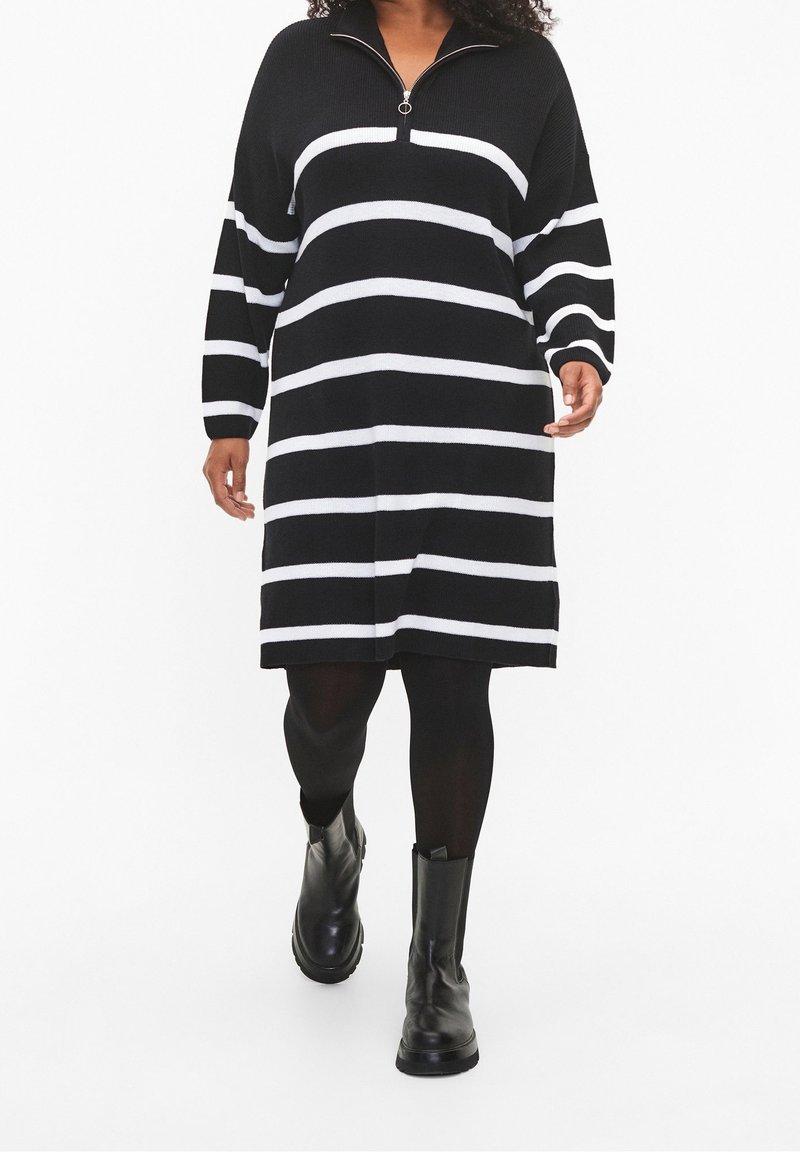 Femme marchant portant une robe noire à rayures horizontales blanches, des collants noirs et des bottines noires sur un fond blanc.