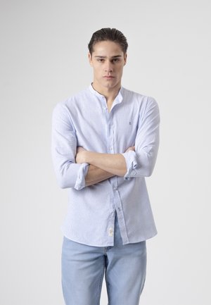 Joven con el pelo peinado hacia atrás, de pie con los brazos cruzados, lleva una camisa azul clara abotonada con las mangas remangadas y unos vaqueros azul claro a juego.