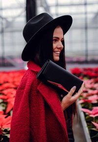 Black leather clutch with a red accent, held against a textured red coat, set among pink floral background.