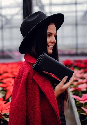 Black clutch with a red accent, textured fabric, and angular design; worn with a bright red coat in a floral setting.