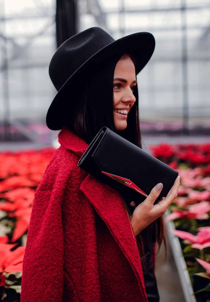 Black leather clutch with a red accent, held against a textured red coat, set among pink floral background.
