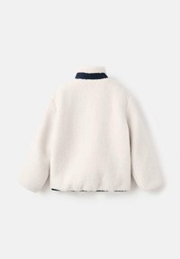 Veste en polaire écrue avec un col marine, dotée d'une texture douce et d'un design court. Manches longues et détails de l'ourlet élastiqué.
