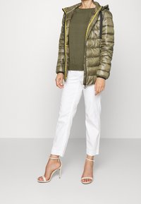 Giacca a vento in verde oliva con zip gialla e dettagli neri, abbinata a un top verde e pantaloni bianchi. Tacchi beige con cinturini completano il look.