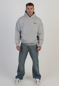 Hanorac gri cu buzunar frontal și logo, asortat cu blugi bleu deschis și adidași albi. Modelul stă în picioare pe un fundal simplu.