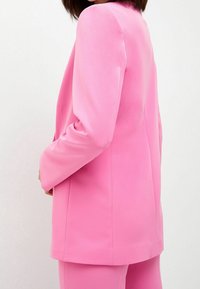 Kobieta w jasnoróżowym żakiecie i dopasowanych spodniach, pokazana od tyłu, stojąca na tle jednolitego białego tła.