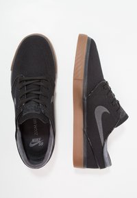 Baskets en toile noires avec un logo Nike gris, dotées d'une semelle en caoutchouc marron et de lacets noirs. Texture lisse avec un design classique de chaussure de skate.