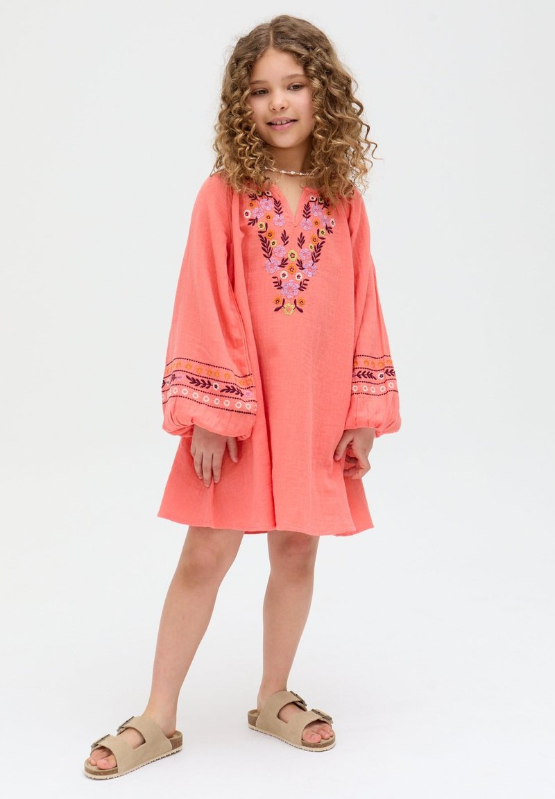 Mädchen mit lockigem Haar, das ein korallenfarbenes Kleid mit Blumenstickerei auf Brust und Ärmeln trägt, steht mit auseinander stehenden Füßen und lächelt.