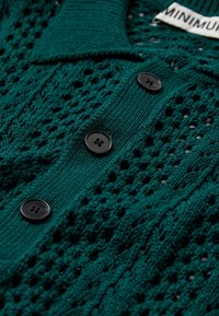 Cardigan en tricot vert foncé avec des boutons noirs et un motif ajouré texturé, montrant une partie d'une étiquette blanche.