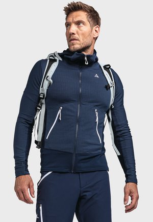 Marineblaue Zip-Jacke aus strukturiertem Stoff, weiße Reißverschlusstaschen und ein hoher Kragen. Kombiniert mit marineblauen Hosen und einem hellgrauen Rucksack.