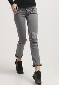 Grå fit jeans i bomullsmaterial, med design av främre fickor och upprullade muddar, kombinerat med svarta träningsskor.