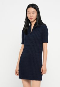 Μπλε πλεκτό φόρεμα με υφασμάτινο σχέδιο, κοντά μανίκια και κολάρο με φερμουάρ. Διαθέτει μια μικρή λογότυπη λεπτομέρεια στο στήθος.