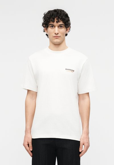 Νεαρός με σγουρά σκούρα μαλλιά, φοράει λευκό μπλουζάκι Barrow και μαύρο παντελόνι, στέκεται μπροστά σε απλό λευκό φόντο.