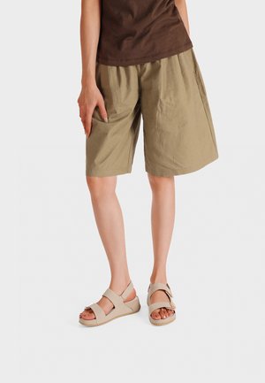 Donna che indossa pantaloncini beige fino al ginocchio, top marrone e sandali beige con punta aperta, in piedi davanti a uno sfondo semplice.