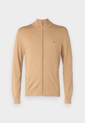 Beige zip-up tröja med hög krage, långa ärmar och en liten logotyp på vänster bröst. Tillverkad av mjukt, lätt material.
