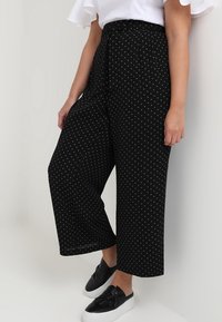 Pantalon noir à jambes larges à pois blancs, avec une taille haute, une ceinture à nouer et un tissu léger. Associé à des chaussures noires sans lacets.