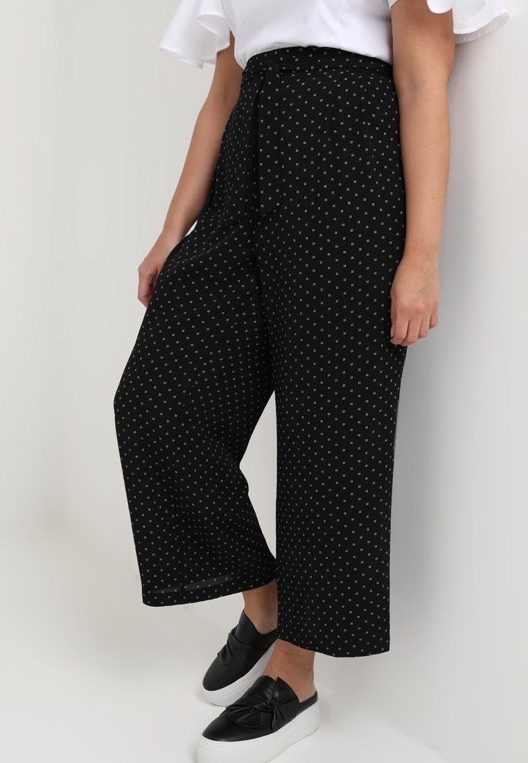 Pantalon noir à jambes larges à pois blancs, avec une taille haute, une ceinture à nouer et un tissu léger. Associé à des chaussures noires sans lacets.