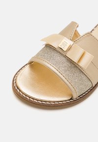 Guld läder sandal med ett glittrande silverbälte, prydd med en satinrosett och metall-logotypdekoration. Slät textur med en rund tådesign.