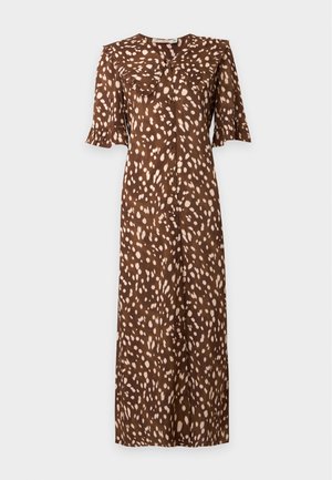 Femme portant une robe midi marron avec des taches claires, col et manches à volants, debout les mains dans le dos et chaussures plates marron.