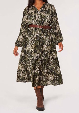 Femme portant une robe verte à manches longues à motif floral, une ceinture marron et des bottines en daim marron sur un fond uni.
