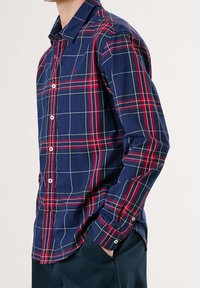 Mężczyzna ubrany w granatową, czerwoną i białą koszulę w kratę z guzikami, z rękawami opuszczonymi i ręką w kieszeni, na jednolitym tle.