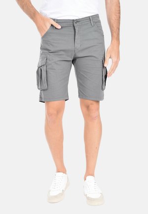 Uomo che indossa pantaloncini cargo grigi con tasche laterali, scarpe da ginnastica bianche e una maglietta bianca, in piedi con una mano in tasca.