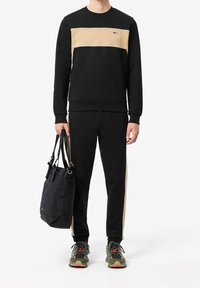 Černá mikina s béžovým panelem, černé tepláky s béžovými bočními pruhy, držení černé tašky a nošení šedých tenisek.