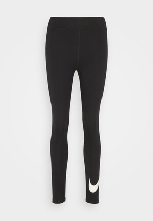 Leggings negros hechos de tela elástica, con una cinturilla alta y un acento de logo blanco en la parte inferior de la pierna izquierda. Textura suave y diseño ajustado.