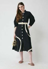 Schwarzes Hemdkleid mit drei Viertelärmeln, cremefarbenen Akzenten und einem gebundenen Gürtel. Getragen mit einer strukturierten Handtasche und flachen Sandalen.