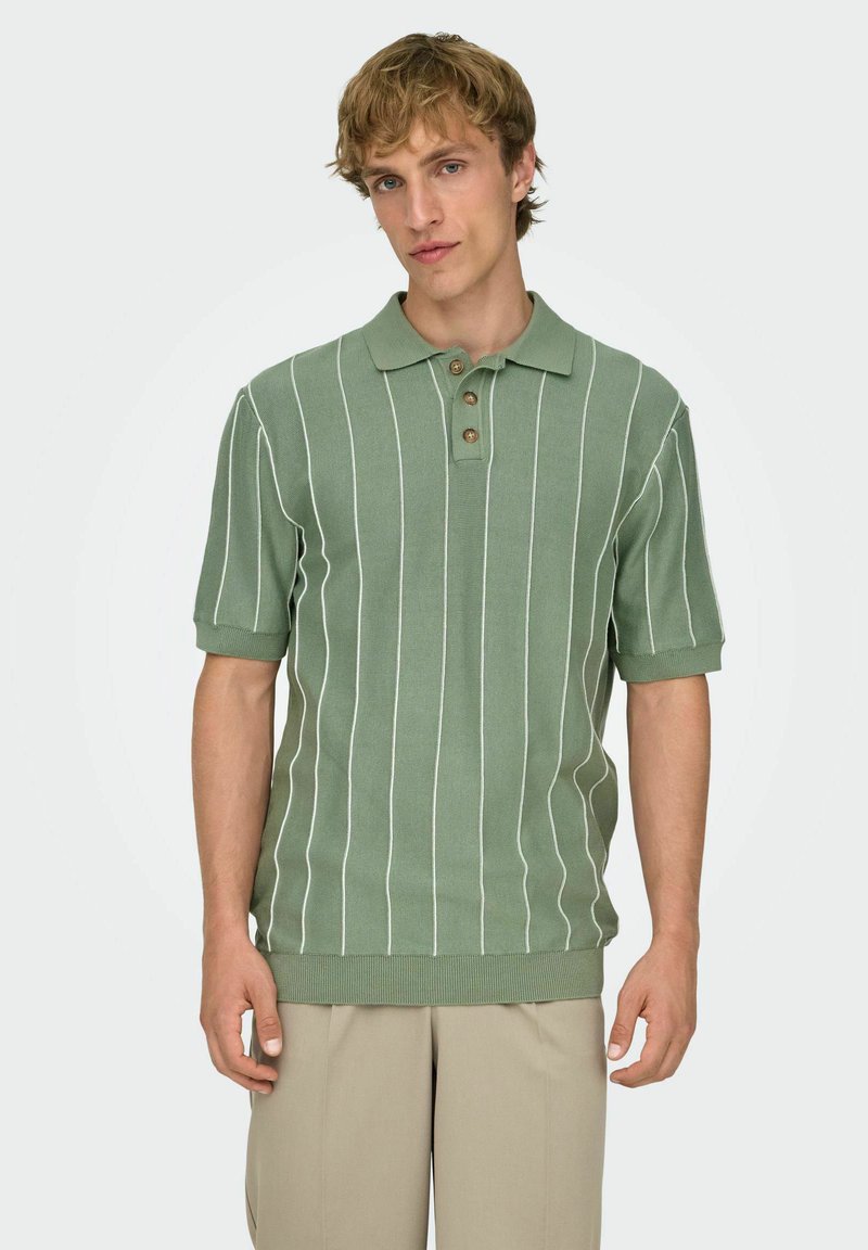 Jeune homme portant un polo vert à manches courtes avec des rayures verticales blanches et un pantalon beige, posant devant un fond clair uni.