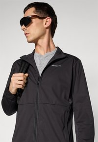 Man wearing black Patagonia jacket, gray shirt, carrying backpack on one shoulder, and wearing black Oakley sunglasses looking left.