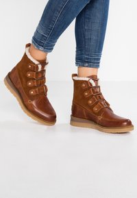 Botas de invierno de cuero marrón y gamuza con forro de piel de pelo, combinadas con jeans azules, sobre un fondo blanco.