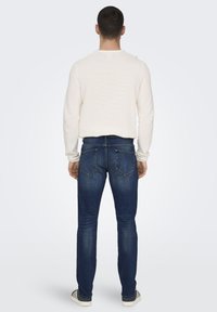 Ein weißer Strickpullover und dunkelblaue Jeans. Der Pullover hat einen Rundhalsausschnitt, während die Jeans ein klassisches 5-Taschen-Design mit sichtbaren Nähten aufweisen.