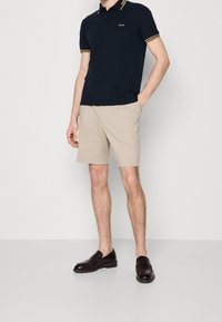 Navy sininen poolopaita, jossa on keltaraidallinen kaulus, beige lyhyet housut ja tummanruskeat nahkasandaalit. Poolopaidassa on logo rinnassa, ja lyhyissä housuissa on laskostettu malli.