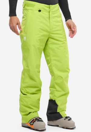 Pantalons de ski vert lime en matériau imperméable, avec une coupe décontractée, des poches zippées et un logo sur la jambe ; portés avec des bottes de ski.