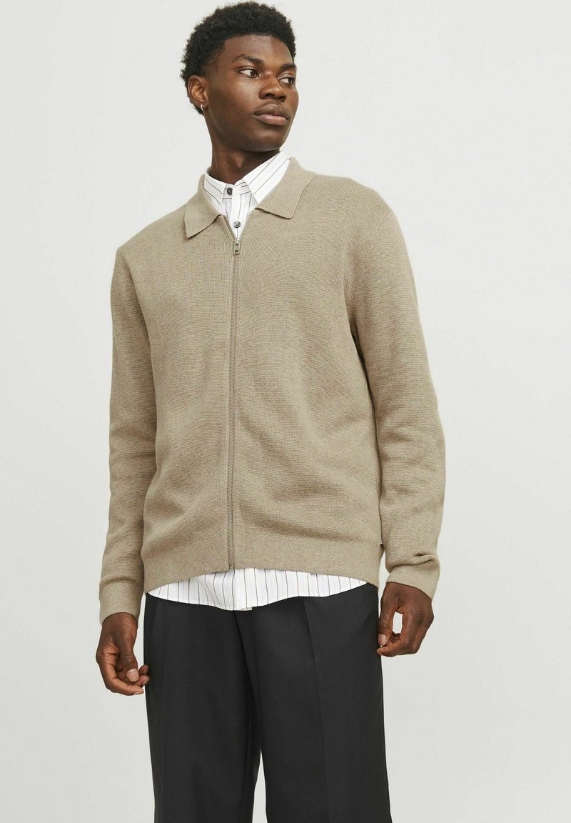 Cardigan beige lavorato a maglia con chiusura a zip, design con colletto, maniche lunghe e finitura leggermente texturizzata, indossato sopra una camicia bianca a righe.
