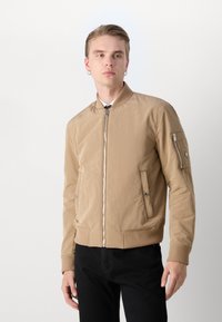 Veste bomber marron clair avec une fermeture éclair à l'avant et des poignets côtelés. Présente des poches latérales et une poche zippée sur le bras. Texture de tissu lisse.
