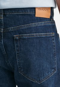 Jeans din denim albastru închis cu o textură netedă, având cusături portocalii contrastante și un patch din piele de culoare bej cu logo-ul GANT pe spate.