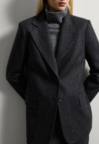 Donkergrijze wollen blazer met een iets gestructureerde afwerking, voorzien van een notch lapel, twee zakken en een enkele knoopsluiting, gedragen over een grijze coltrui.