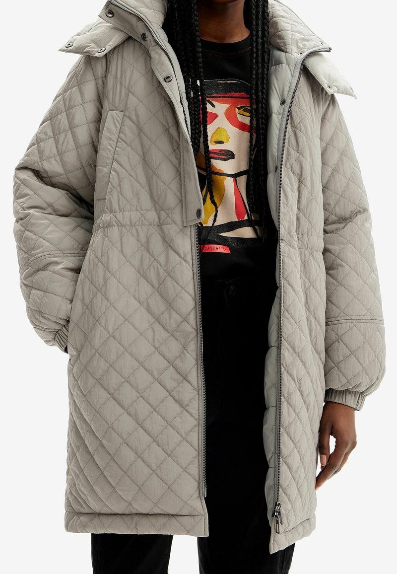 Personne portant un long manteau matelassé beige sur une chemise noire ornée d'un visage coloré et abstrait, mains partiellement visibles.