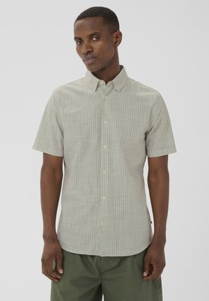 Mand iført en lys beige kortærmet skjorte med subtilt ternet mønster og olivengrønne bukser foran en ensfarvet baggrund.