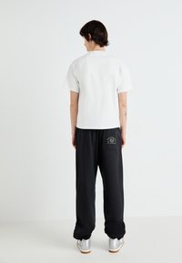 Tricou alb cu mânecă scurtă, asortat cu pantaloni scurți negri din material moale, ce au o buzunar cu logo. Modelul poartă pantofi argintii. Fundal simplu.