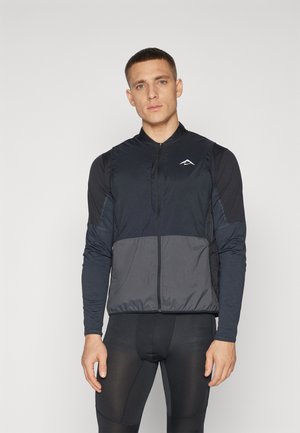Chaleco negro sin mangas con cremallera frontal y panel gris, superpuesto a una camiseta de manga larga azul marino. Presenta un emblema en el pecho.