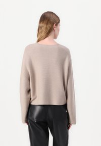 Beige kötött pulóver széles, kerek nyakkivágással és ejtett vállakkal, fekete bőrnadrággal párosítva, hátulról nézve.