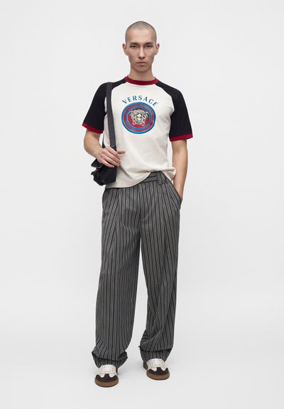 Νεαρός άνδρας που φοράει λευκό και μαύρο t-shirt Versace, γκρι παντελόνι με ρίγες, αθλητικά παπούτσια και μαύρη τσάντα ώμου σε απλό λευκό φόντο.