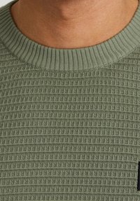 Zielony, fakturowany sweter dziany, z prążkowanym kołnierzem i małą czarną metką z boku. Tkanina ma subtelny, wypukły wzór.